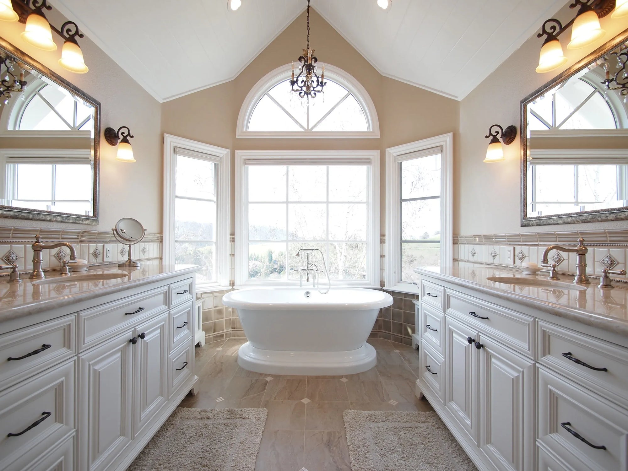 Luxury bathroom with freestanding tub, arched garden windows, dual marble vanities, brass fixtures and chandelier lighting.