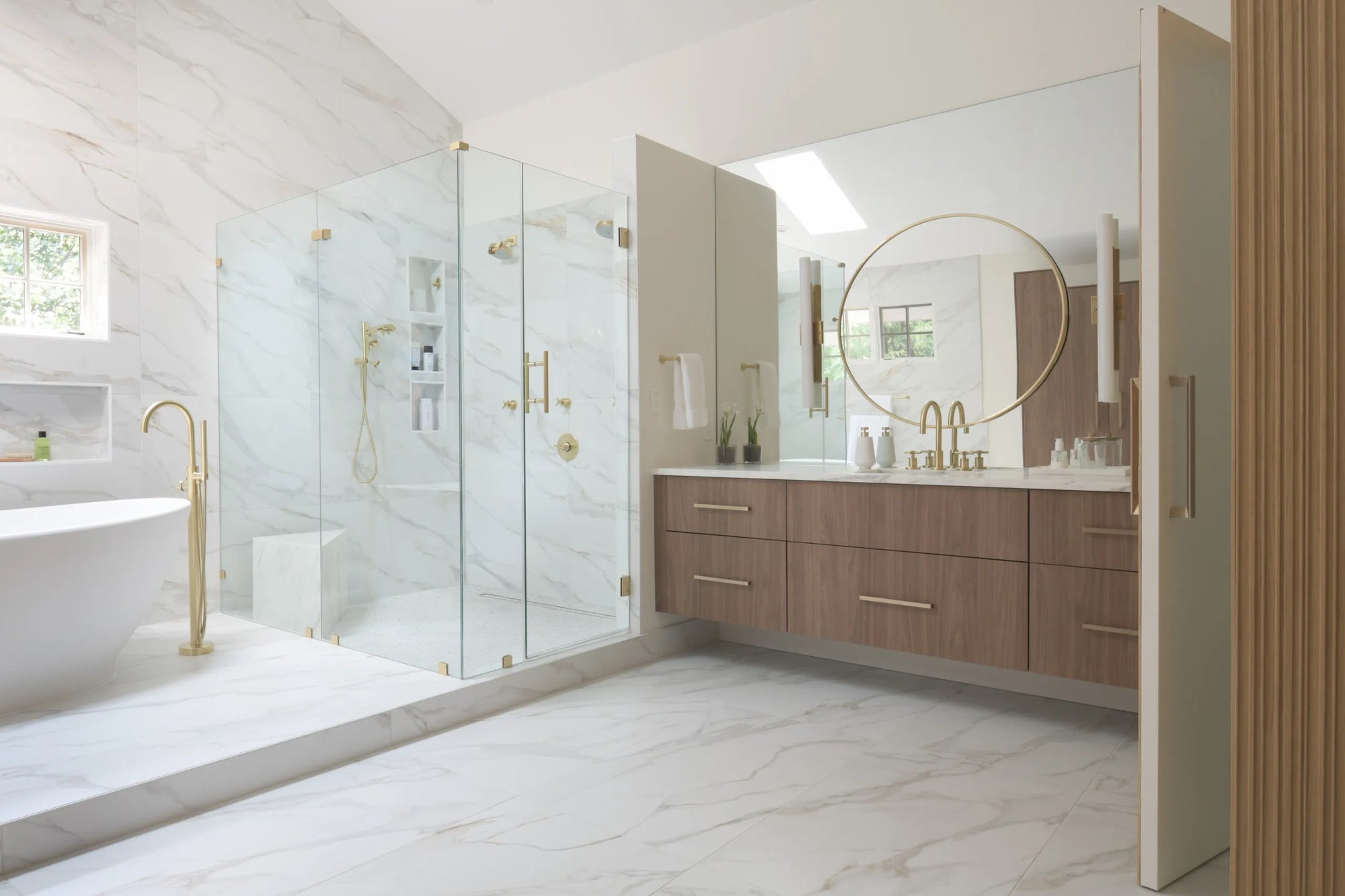 Frameless glass shower door with brushed gold hardware in marble bathroom spa retreat.​