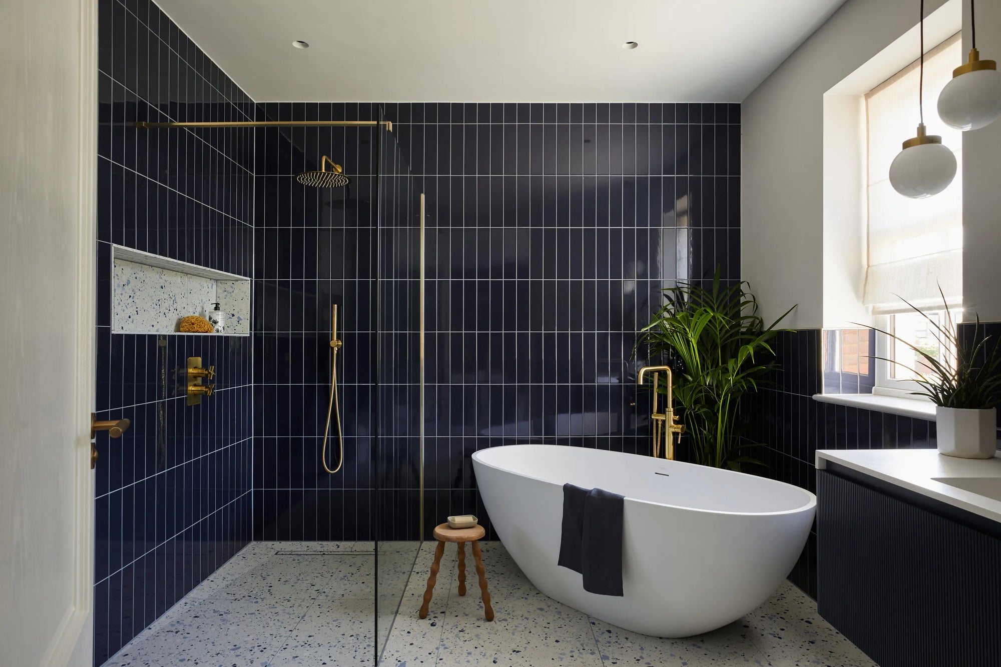 Modern bathroom with glass shower door, dark blue tiles, freestanding tub, gold fixtures, and natural light. Stylish & luxurious.