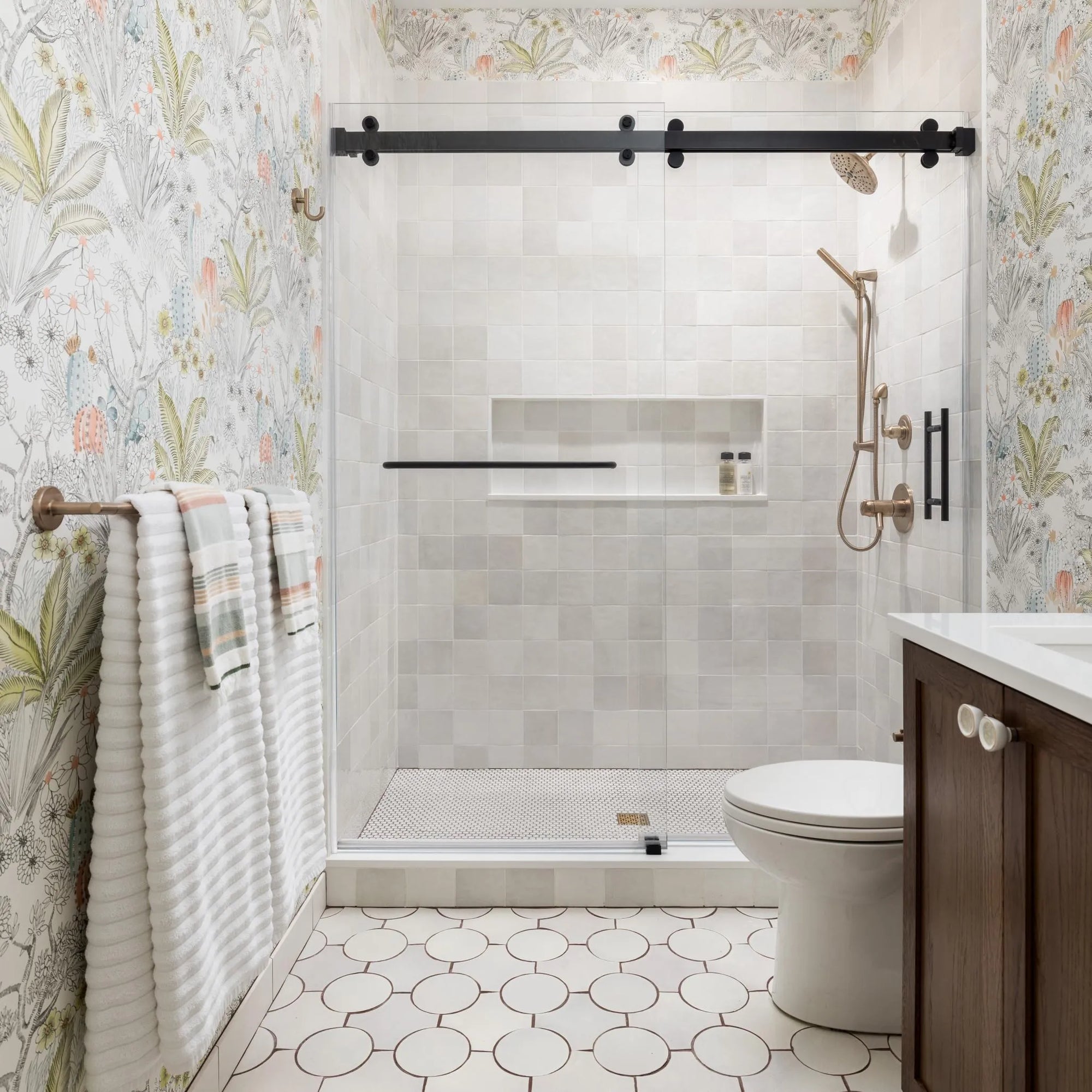 Modern walk-in shower with a black barn door, floral wallpaper, and a rose gold showerhead against grey subway tiles.