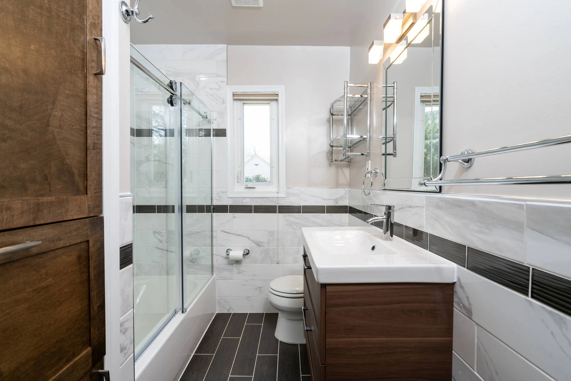 Glass shower door enclosing bathtub-shower combo with dark wood vanity, white sink, chrome fixtures in modern bathroom.