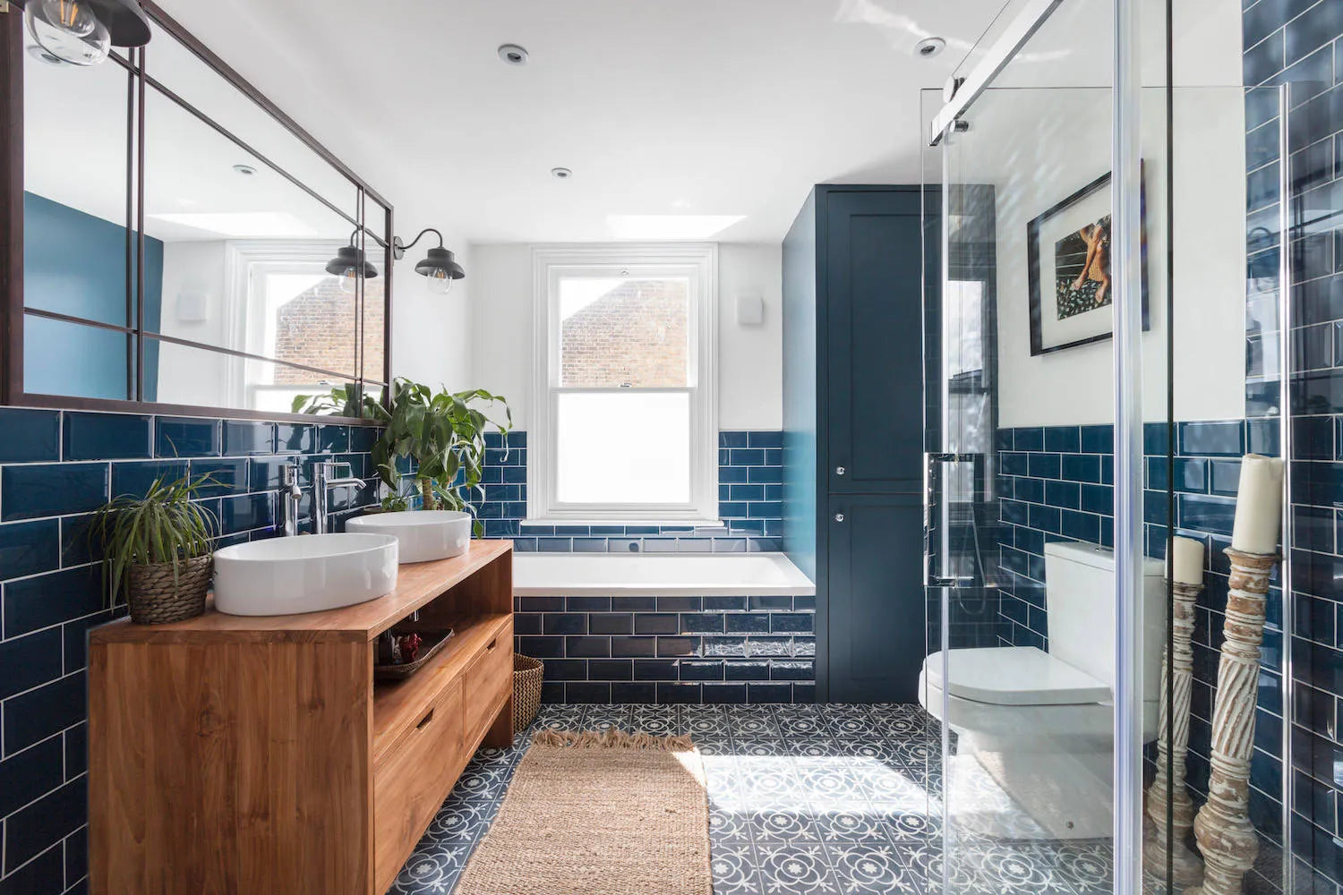 Modern bathroom with frameless shower door, blue subway tiles, vessel sinks, wood vanity, patterned flooring, and freestanding tub.​