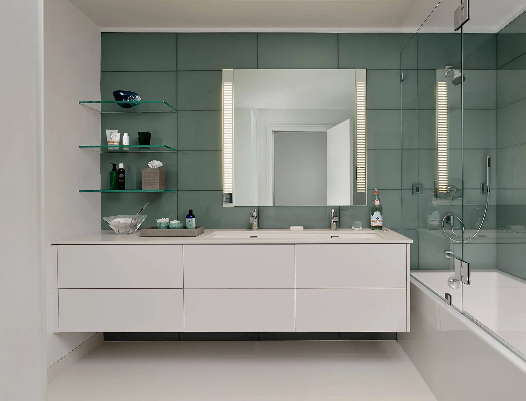 Modern bathroom with floating double vanity, LED-lit mirror, and transparent floating glass shelves against green tile wall.​