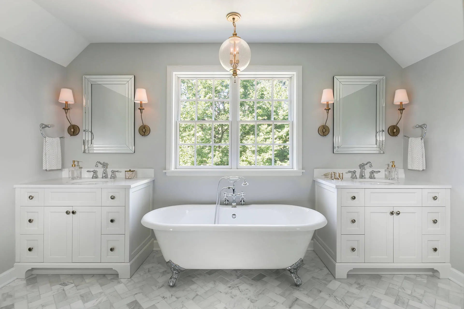 Freestanding clawfoot bathtub flanked by double vanities in elegant symmetrical bathroom with marble flooring and large grid window.