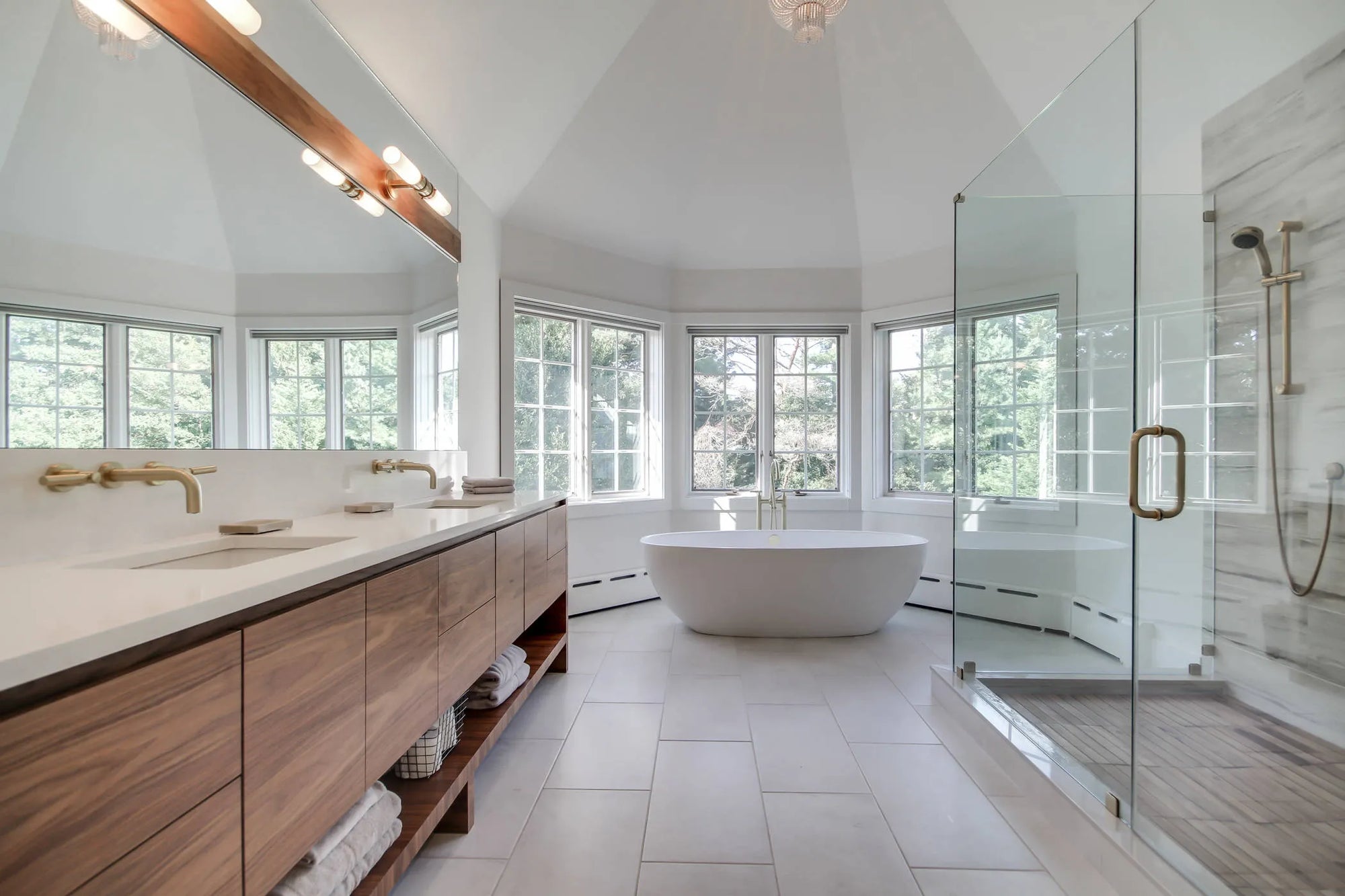 A sleek white freestanding bathtub in a luxurious modern bathroom with large windows, gold fixtures, and vaulted ceilings.