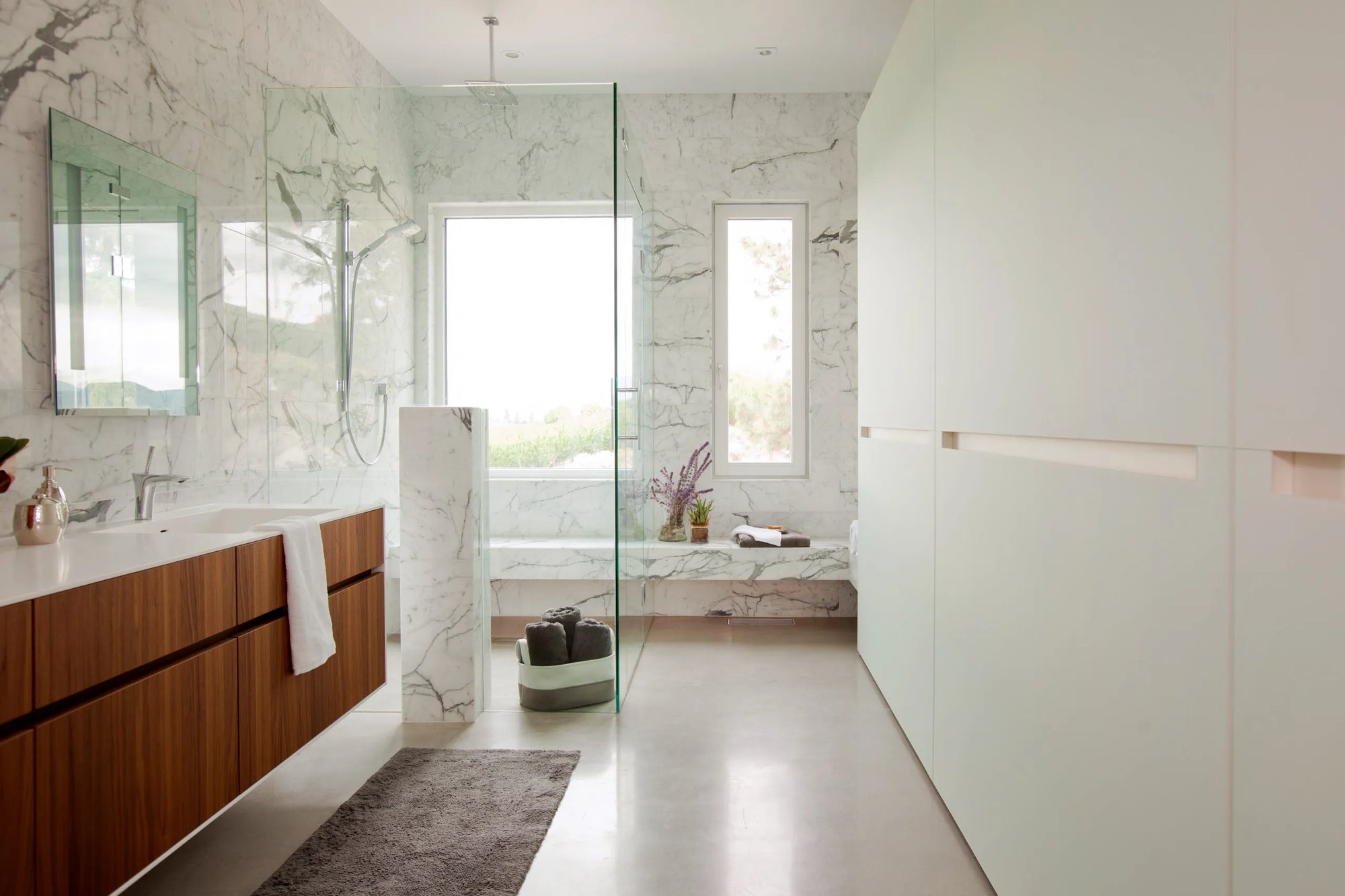 Modern minimalist bathroom with bathroom bench near window, marble walls, wood vanity & glass shower partition.​