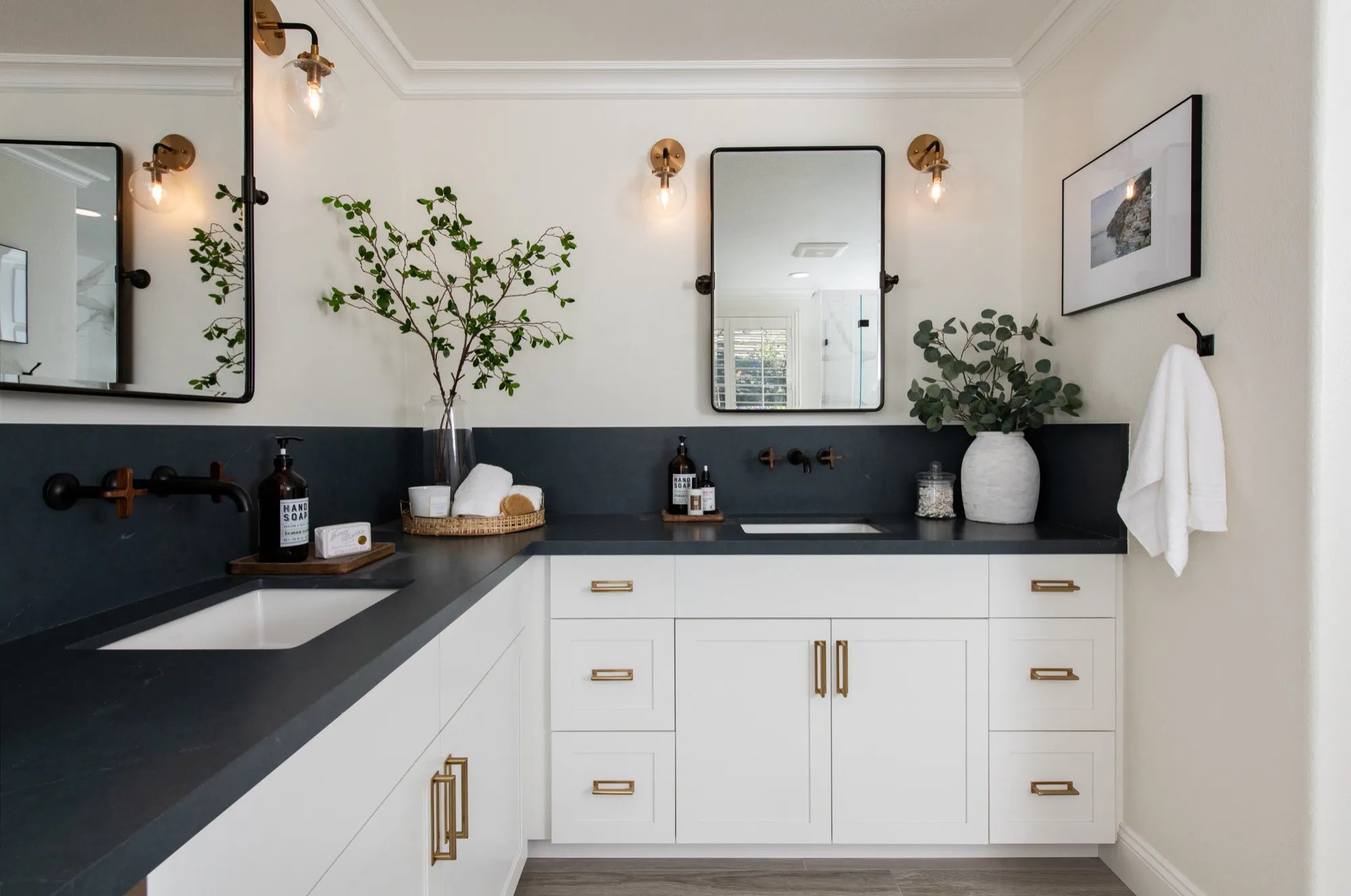 Modern L-shaped bathroom vanity with white cabinets, gold fixtures, dark countertop, and double sinks.