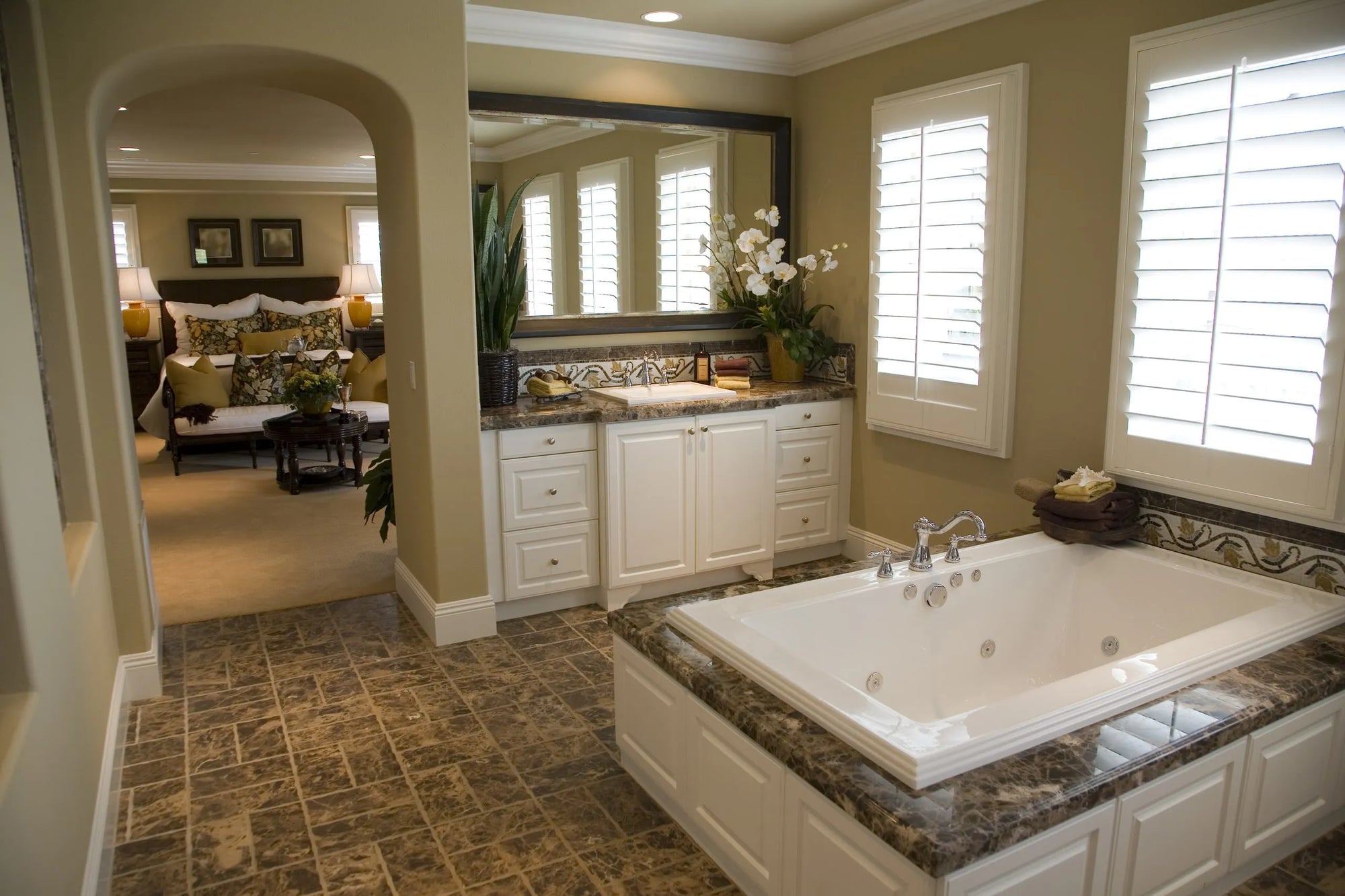 Luxury bathroom with a Jacuzzi bathtub under large windows, featuring a double vanity and an arched doorway to an adjacent bedroom.