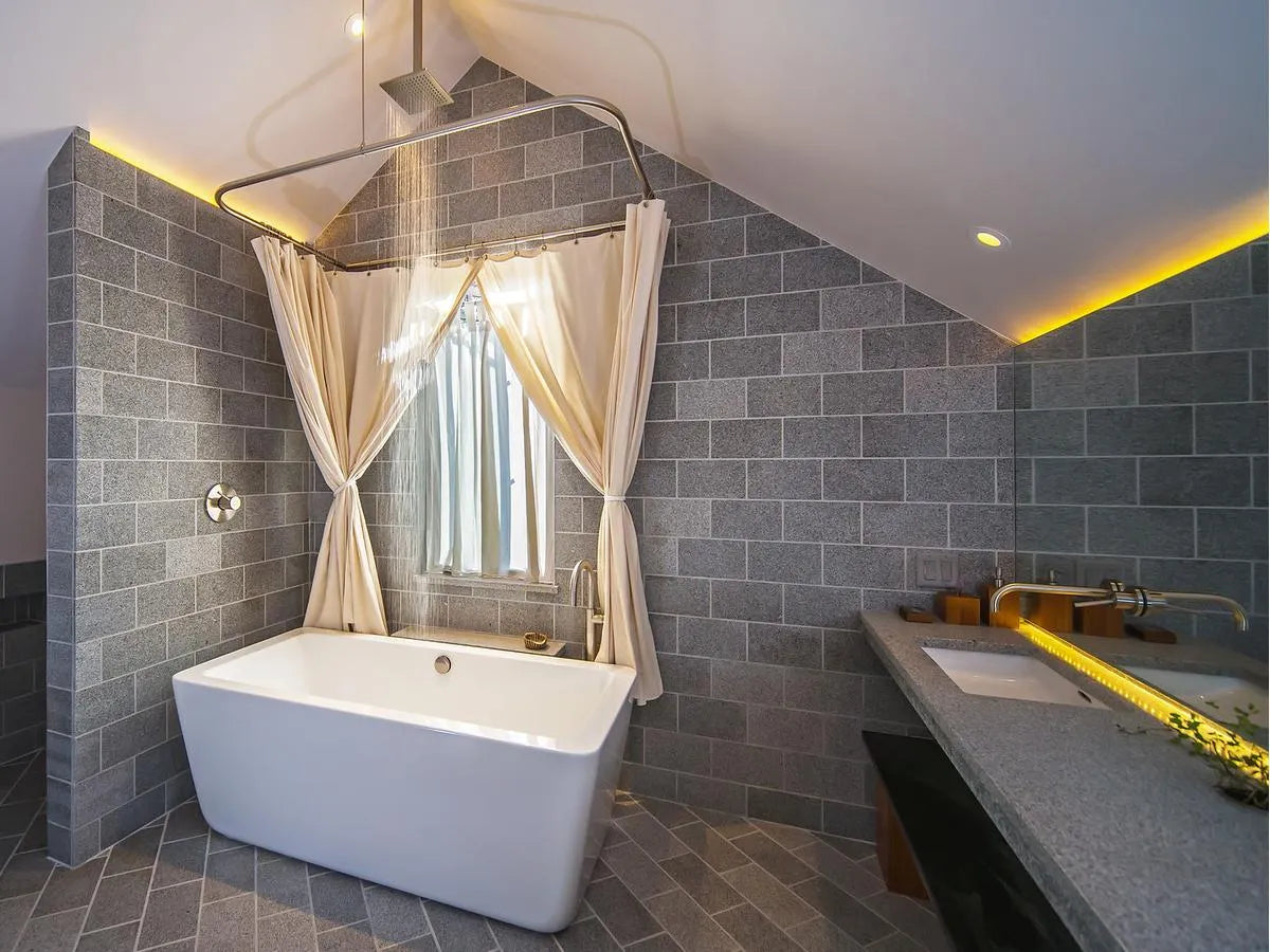 Luxurious bathroom with a freestanding tub with shower under a rainfall showerhead, gray brick walls, gold fixtures, and a slanted ceiling with warm lighting.