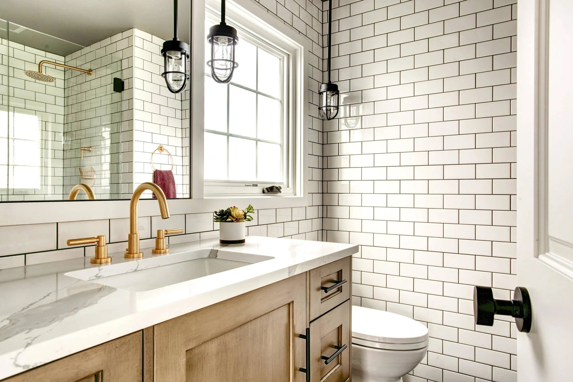 Stylish modern farmhouse bathroom with white subway tiles, a gold faucet, a elegant vanity and black industrial pendant lights.