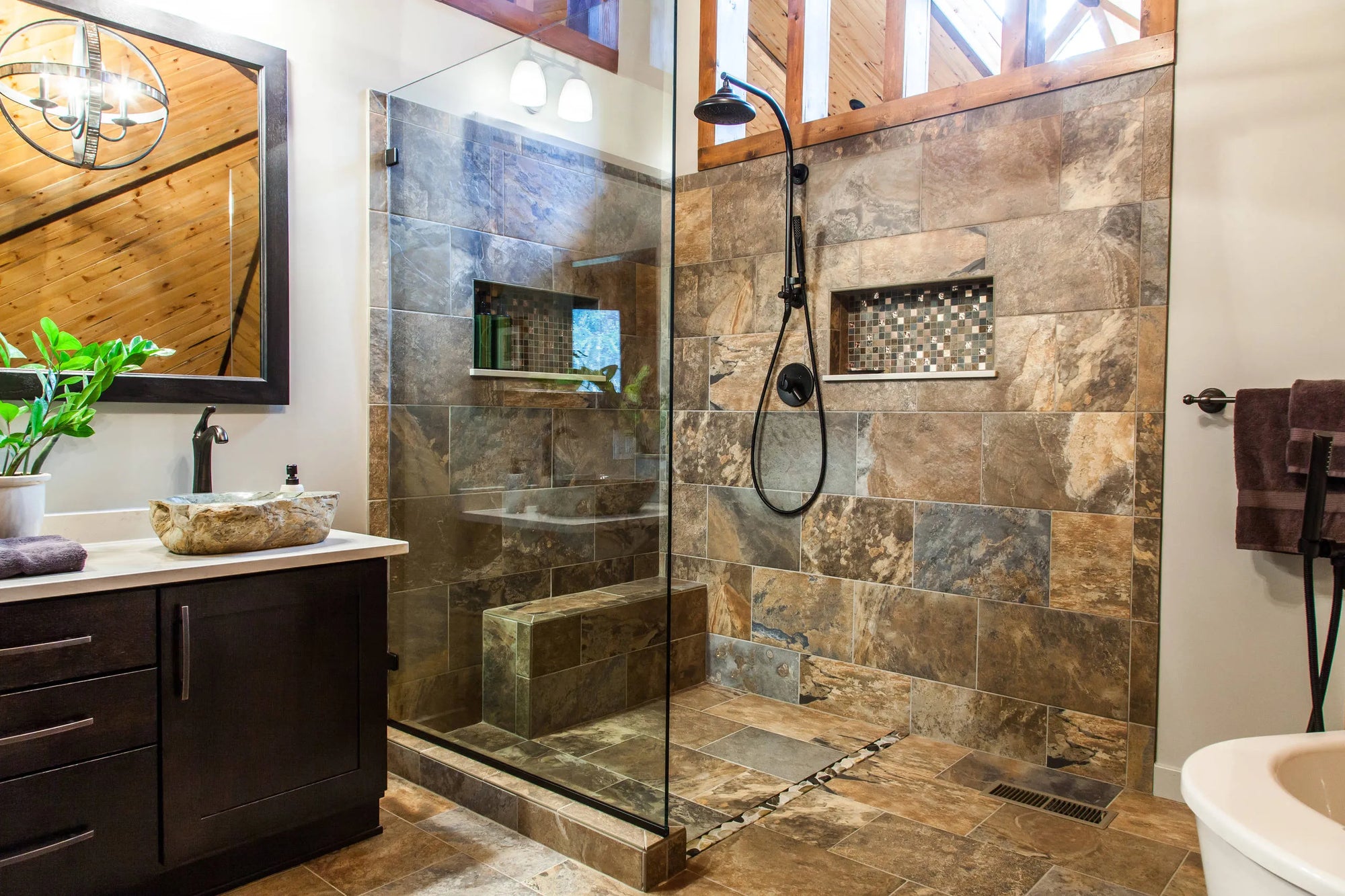 Luxurious bathroom with Venetian Bronze fixtures, natural stone tiles, glass shower, and wooden beams.