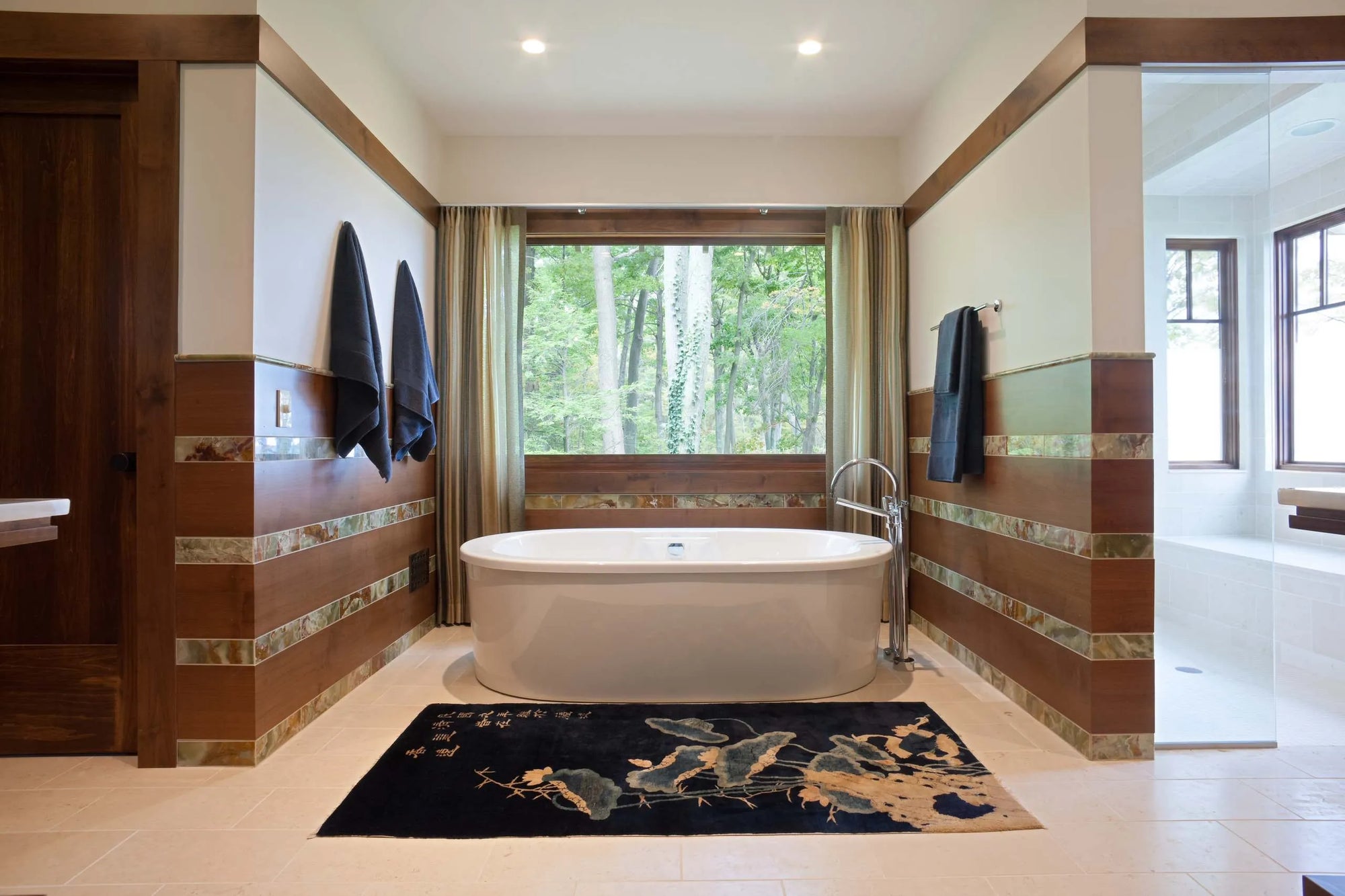 Spa-like master bathroom featuring a freestanding bathtub by forest-view windows, with warm wood accents, decorative tile, and plush rug for a luxurious retreat ambiance.