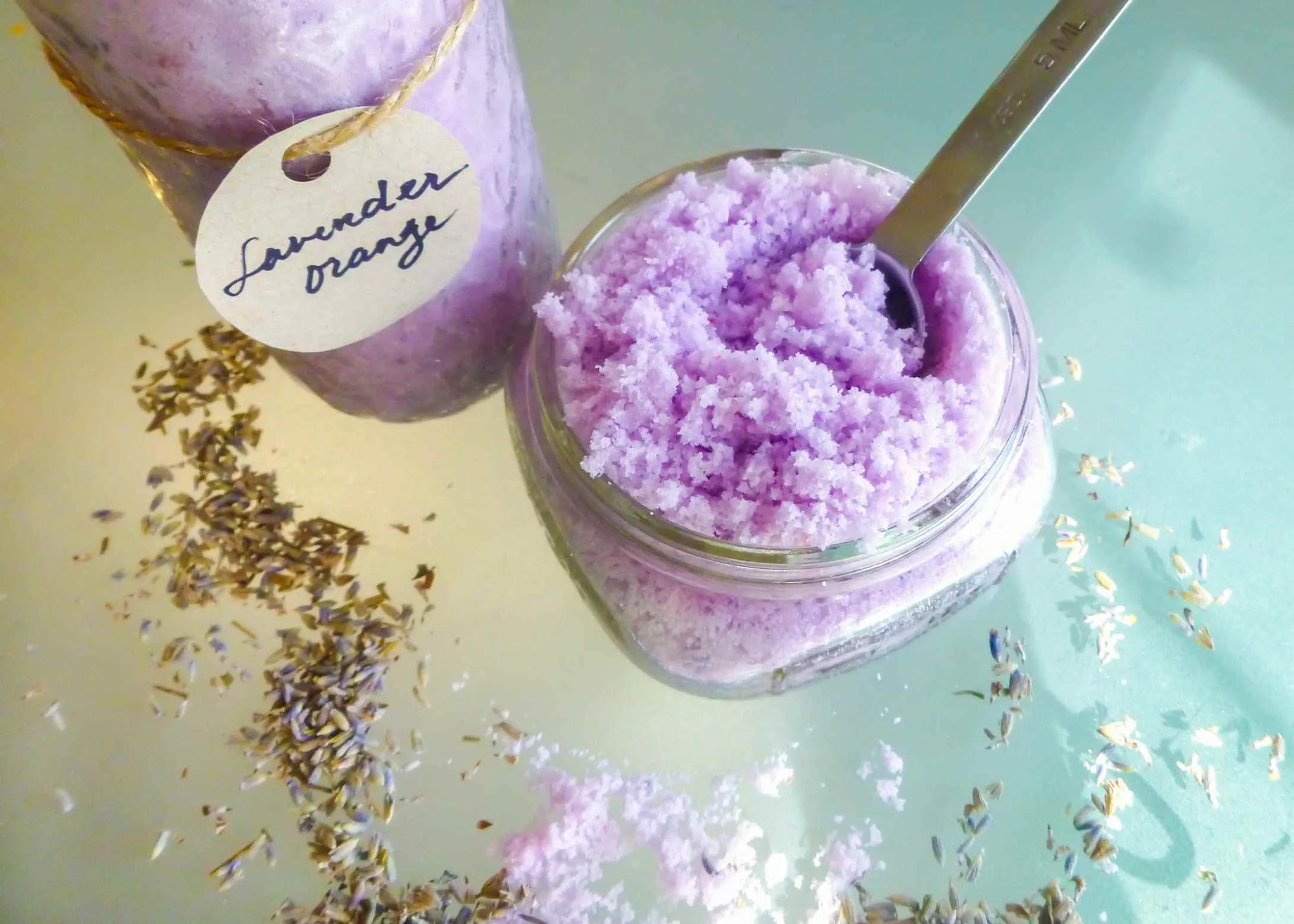 Purple bath salts in glass jars with spoon, labeled "Lavender Orange," scattered texture on light surface, soft spa aesthetic, minimalist bathroom product display.