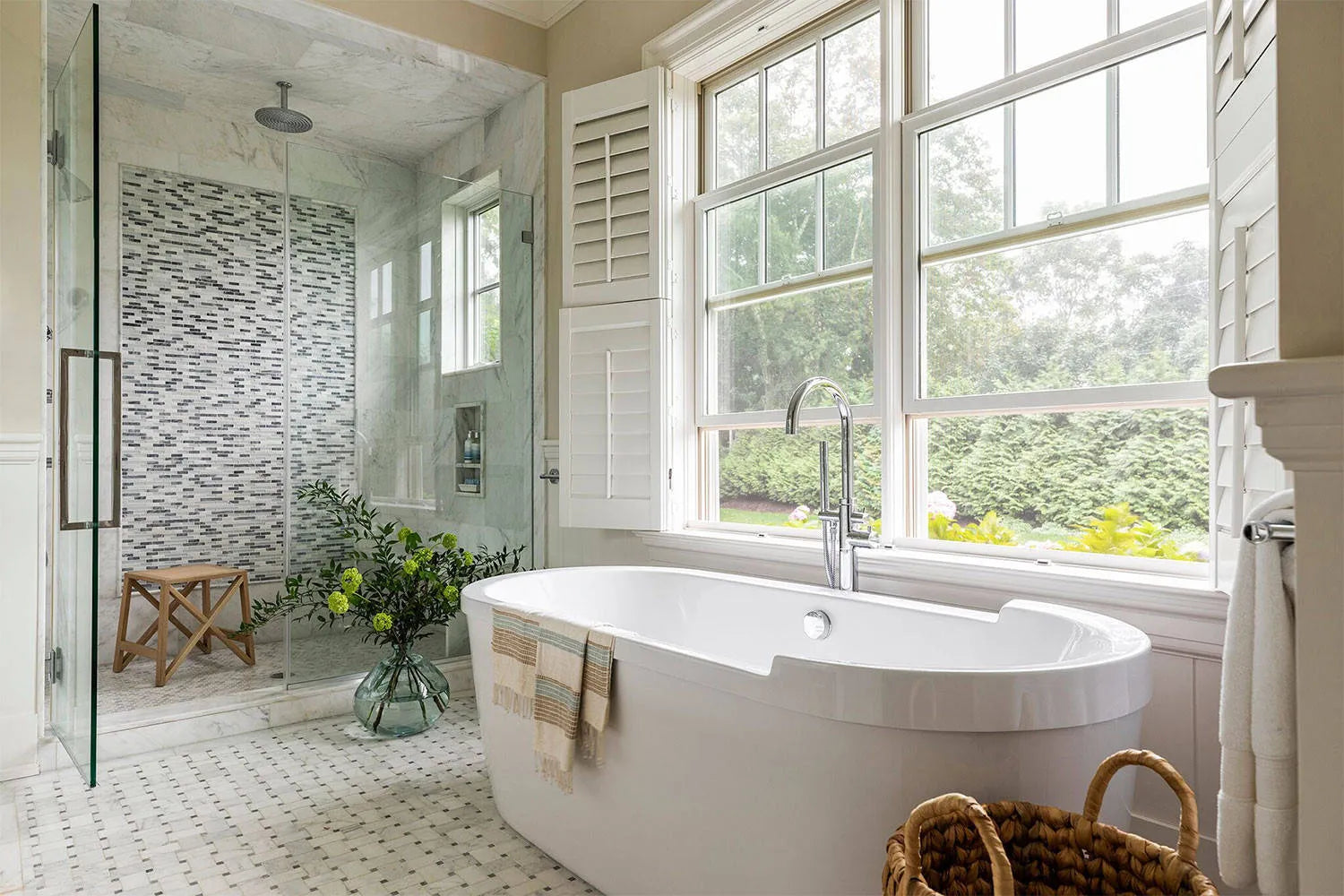 Bright bathroom with freestanding bathtub, frameless glass shower, and large windows overlooking greenery.