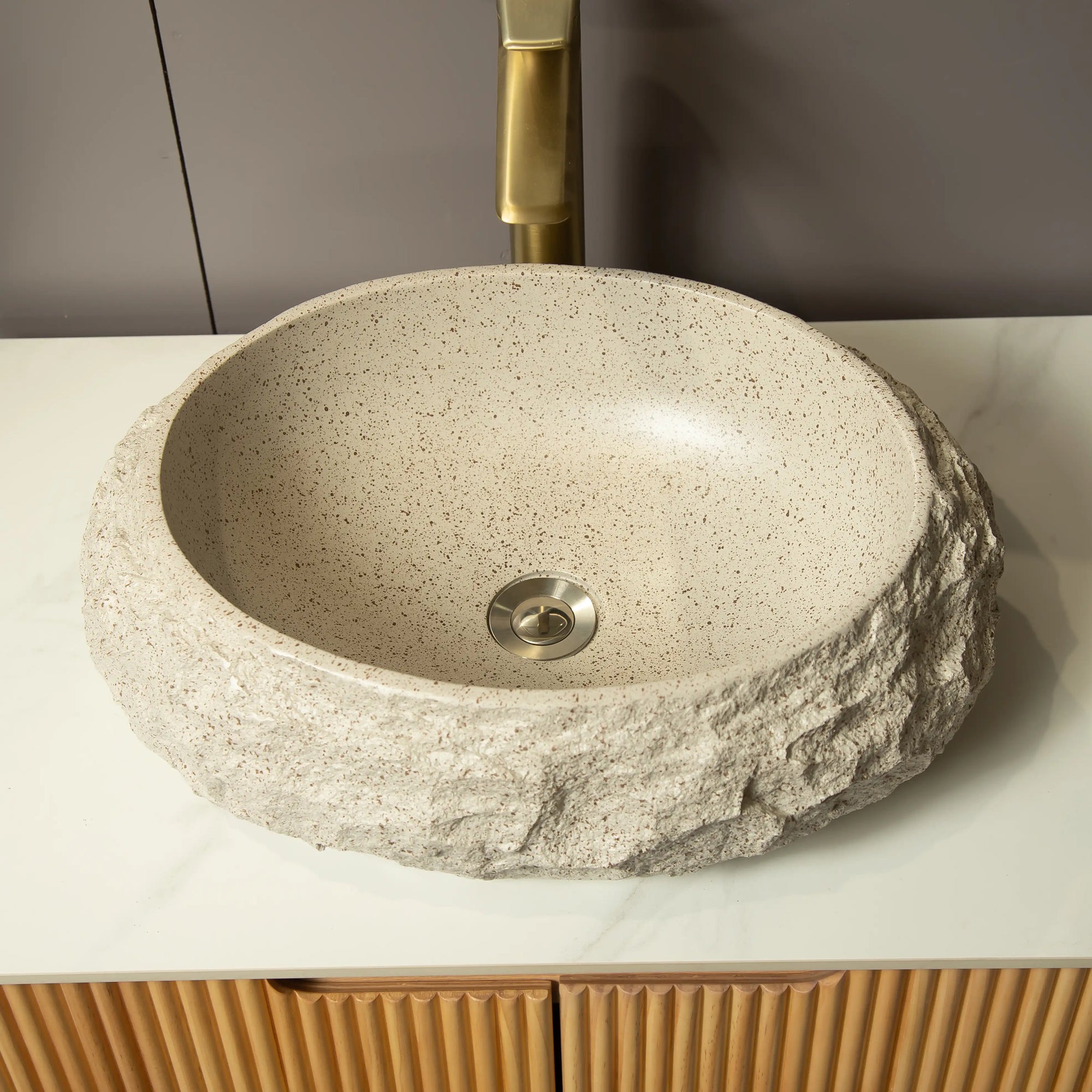 A beige stone-textured vessel sink with a rough exterior and smooth interior, set on a white countertop beneath a brass faucet. |ACE DECOR