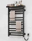 Black electric towel warmer with top storage shelf and LED touch controls mounted on white bathroom wall.