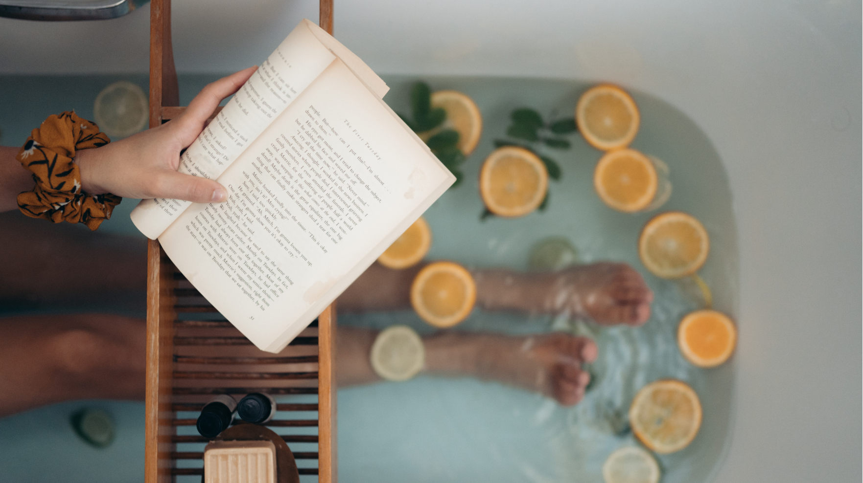 A person holding an open book, with feet in the water of their bathtub filled with lemon and orange slices, seen from above. | ACE DECOR