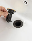 Hand installing a black and gold drain stopper into a white sink, compatible with waterfall faucet bathroom setups | Ace Decor